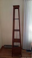 Full view of the tall wooden narrow shelf unit against a wall, showing tapering form and four shelves plus top shelf.