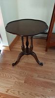 Photo of dark wood pedestal side table showing scalloped edge on tabletop, three turned supports, and three decorative carved legs on hardwood floor.