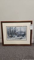 Full view of the framed print depicting deer in a winter woodland setting with a rustic cabin, showing the frame and matting, on grey carpet against a white wall