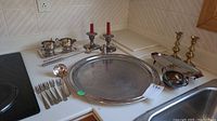 Full view of all silver plated items including tray, forks, ladle, sugar bowl, creamer, candle sticks, and snuffer arranged on a kitchen counter