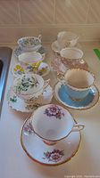 Set of eight teacups and matching saucers on a white surface showing various floral patterns and gold detailing.