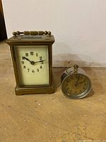 Two vintage clocks on a surface: rectangular brass carriage clock and small round silver-tone alarm clock.