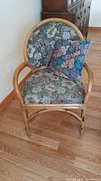 Front angle of wicker chair with floral patterned upholstery and paisley throw cushion showing seat and rounded backrest.