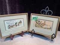 Two signed prints in gilded wood frames shown on stands against black background