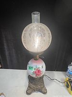 Full view of the antique Gone With The Wind style lamp with frosted etched glass shade, rose painted globe base and ornate cast iron base on table with power cord and water bottle visible.