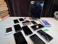 Lot of cell phones including an iPad, several smartphones with cracked and cracked screens, older slide-out keyboard phones, and a new Apple phone case in packaging. All displayed on a white surface with books in the background.