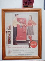 Wood framed 1955 Coca-Cola advertisement depicting a red Coca-Cola vending machine with a man handing a bottle of Coke to a woman, showing overall condition and size.