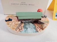 Top view of the Country Covered Bridge figurine showing the green roof, red sides, and textured river landscape base with painted details.