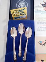 Three silverplate presidential spoons resting in blue velvet case with black outer case, showing the front embossed portraits on handles and presidential seal on case lid.