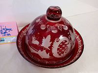 Front view of red cranberry glass covered dish showing full dome lid with etched nature motifs including deer, leaves, swirls, and patterned elements, placed on round plate base with matching red cranberry glass.