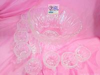 Complete set of clear vintage square cut glass punch bowl with 8 matching cups arranged around it on a pink background.