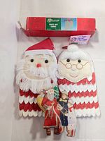 Two fabric Christmas pot holders featuring Santa Claus and Mrs. Claus faces with red and white layered fabric and faux fur details, and a hanging Santa scene ornament with vintage-style embroidery.