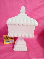 Front view of the 1950's Westmoreland square pedestal milk glass dish with lid showing geometric patterns and finial handle.