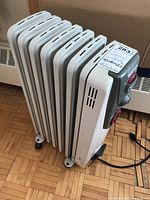 Side view of DeLonghi Magnum MG-15T space heater showing white fins, mobility wheels, and power cord on wood parquet floor.
