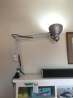 Full view of adjustable metal desk lamp with clamp base and rounded bowl-shaped shade, attached to a white desk.