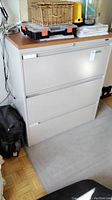 Front view of Global 3-drawer lateral file cabinet with wood grain top, off-white metal body, closed drawers, and items on top including a wicker basket and tool box.