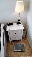 Photo of the two tabletop lamps on either side of a white nightstand with a black clock radio on top and healthometer scale on the floor.