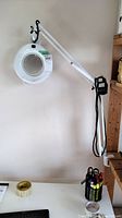 Full view of the clamp-on magnifier flex lamp mounted on a wall above a desk, showing the arm, lens, and clamp base.