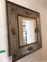 Square mirror with metal frame showing embossed gold-tone floral and abstract decorations from a side angle. Wall and kitchen background.