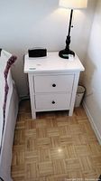 White Ikea Hemnes nightstand next to a bed with lamp and clock on top