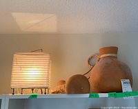 Small square paper and metal lamp lit and shown on white shelves with gourd vase and jug next to it.
