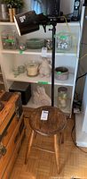 Photo of black metal floor lamp next to round wooden stool placed on floor in front of shelves.