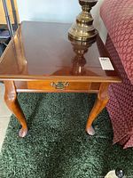 Top and side view of one wooden end table showing drawer, its metal pull handle, and Queen Anne style legs on carpet.