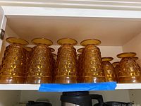 Six amber pressed glass tumblers displayed upside down on a shelf, showing the textured geometric pattern and flared bases.