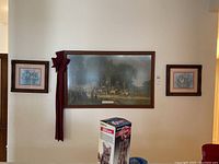 Three framed art pieces mounted on a wall: two smaller floral prints on the sides and one large Fourth of July Parade print in the center with a maroon bow attached.
