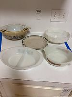 Photo showing six vintage bakeware and mixing bowls arranged on a kitchen counter including divided Pyrex dish, green floral oval dish, scalloped pie dish, stoneware plate, and two floral patterned large mixing bowls.