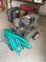 Full view of vintage Briggs & Stratton air compressor on wheels with attached green hose coiled around front, showing overall condition and size relative to surrounding items.