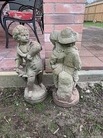 Pair of two concrete garden statues placed on soil next to brick patio, showing full body of each statue from side and back angles.