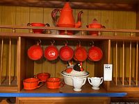 Full shelf view showing orange ceramic beverage set with coffee pot, sugar bowl, six cups, creamer, and white ceramic rooster serving dish along with milk glass creamer and sugar bowl.