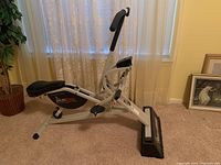 Side view of Rider Riser exerciser showing black cushioned seat, white metal frame, and black base with 'Rider Riser' branding.