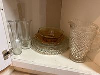 Glassware including clear and amber bowls, glass serving trays, and a large pitcher with diamond cut pattern, displayed on a shelf.