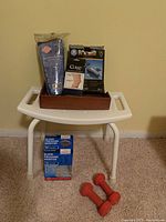 White plastic shower bench with handles, wood tray holding small personal care items, boxed blood pressure monitor, and orange hand weights on carpeted floor