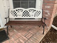 Front view of iron and wood garden bench placed on brick patio. Shows white floral design on backrest, wooden slats for seating, and iron armrests and legs.
