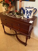 Front and side angle view of the wooden console table with items on top and visible drawer fronts and decorated legs.