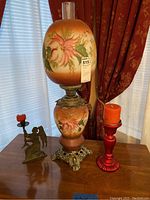 Photo showing one metal and glass globe lamp with floral design, and two candle holders with candles on a wooden table.