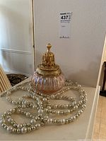 Overall photo showing the pink luster glass oil lamp with ornate gold-tone lid and decorative strand of faux pearl beads placed around the lamp
