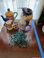 Overview of all the pottery and decorative items on a dark wooden table including the colorful teapot, containers, pottery vase, glass vase and decorative plate along with a string of green beads.