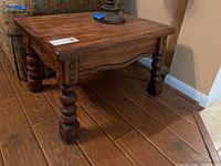 Side view of wooden side table showing detailed turned legs and side panel with carved accents, placed on wood flooring next to upholstered furniture.