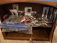 Shelf with various hand tools and supplies in original packaging including scissors, screwdrivers, knives, and other hand tools.