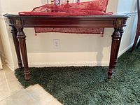 Front view of dark wood console table with carved legs and gold-colored metal accents on corners, placed against a wall on green carpet.
