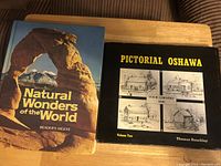 Both coffee table books shown on a wooden surface, one titled 'Natural Wonders of the World' and the other 'Pictorial Oshawa Volume Two' with sketches on the cover.