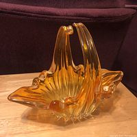 Amber art glass vase shaped like a basket with ruffled edges and arching handle, sitting on wood surface from side angle.