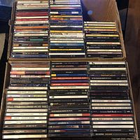 View of stacked CDs in jewel cases, arranged upright showing spines with titles and artists, mostly greatest hits and popular albums.