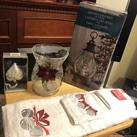Photo showing all lot items on a wooden surface including the glass candle holder, holiday lantern box, towel set, and ornament box.