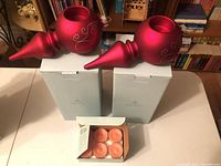 Two red Partylite ornament-shaped tealight holders displayed on white boxes along with an open box of peach-colored tealights on the floor in front.
