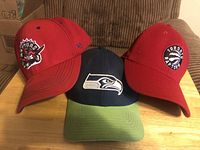 Front view of three sports caps: two Toronto Raptors red caps and one Seattle Seahawks cap.